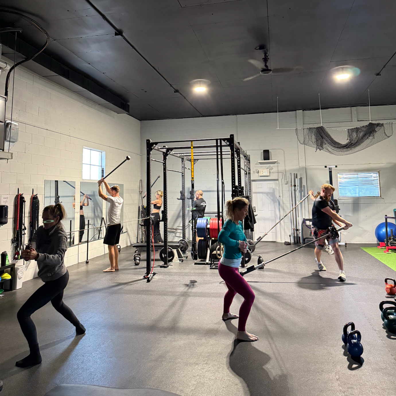 Group of six people exercising with resistance bands attached to a central rack in a gym with kettlebells and weight plates.