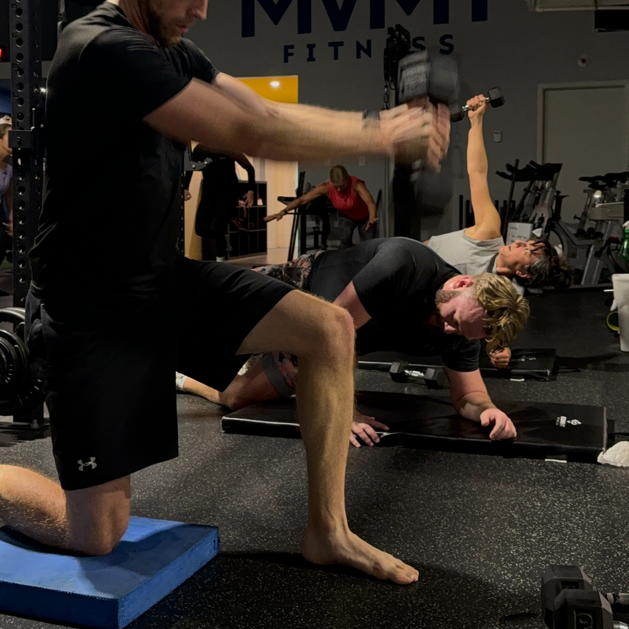 People exercising with dumbbells and mats in a gym with MVMT Fitness signage on the wall.
