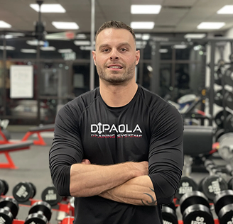 A man with a muscular build stands confidently in a gym, wearing a black "Dipaola Training Systems" shirt. Exercise equipment is visible in the background.