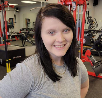 Smiling woman with dark hair and gray shirt in a gym, surrounded by red and black exercise equipment, conveying a positive and energetic mood.