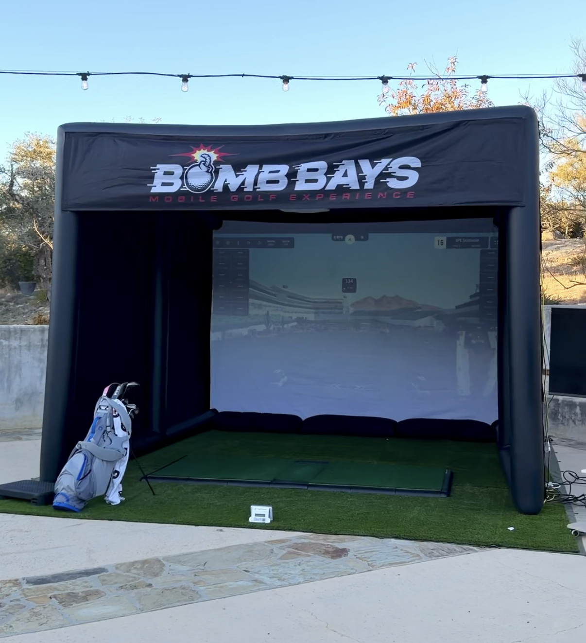 Outdoor mobile golf simulator setup with a large black inflatable enclosure labeled Bomb Bays Mobile Golf Experience, golf bag with clubs on the left, and a virtual golf course projected on the screen inside.