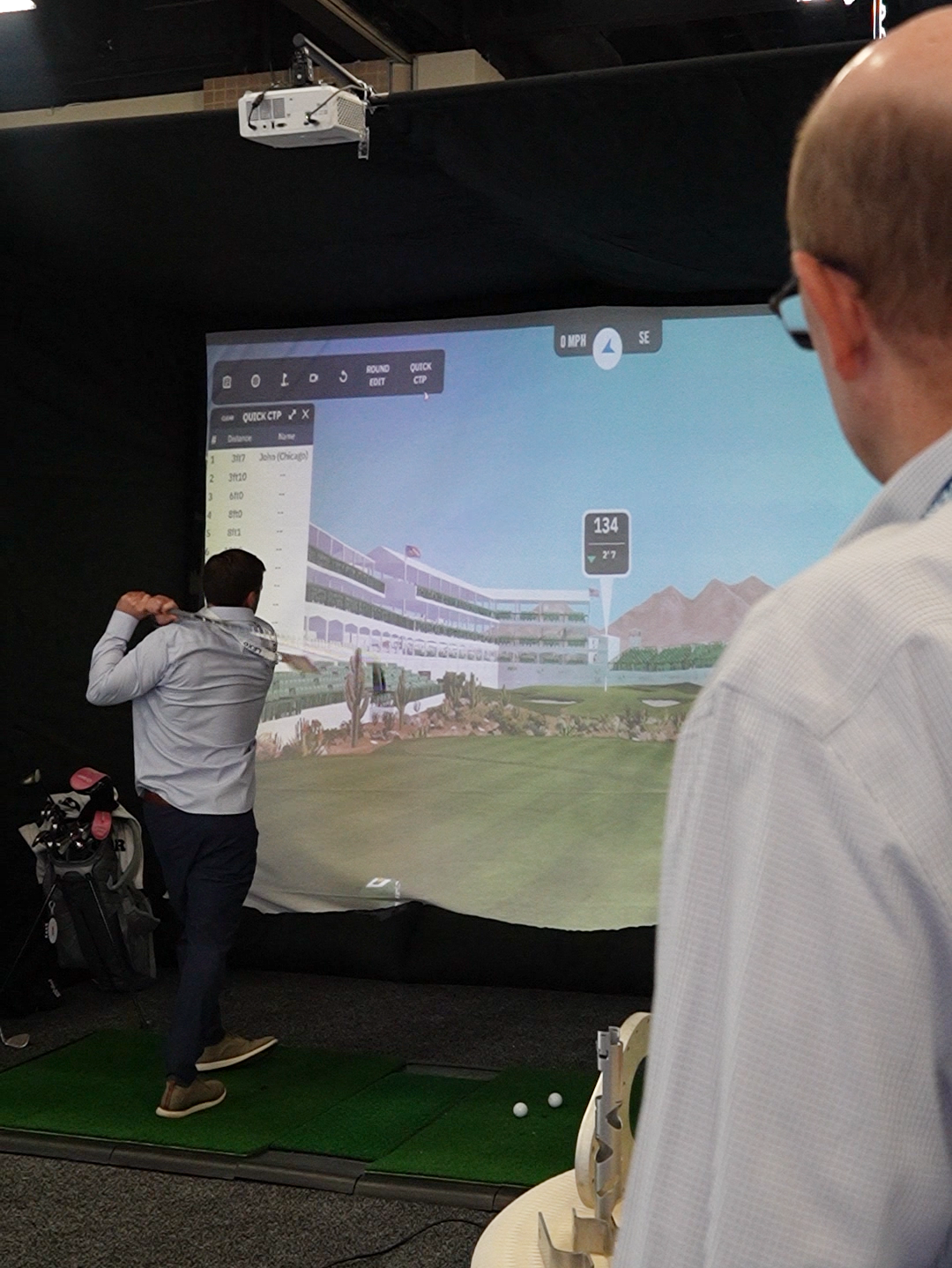 Man swinging a golf club on an indoor golf simulator while another person watches.