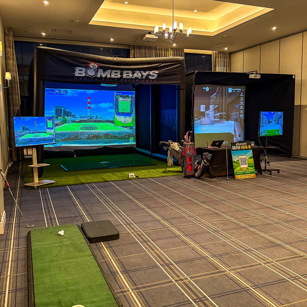 Indoor setup of two Bomb Bays golf simulators with screens showing golf courses, golf bags, and a sign for golfer check-in.