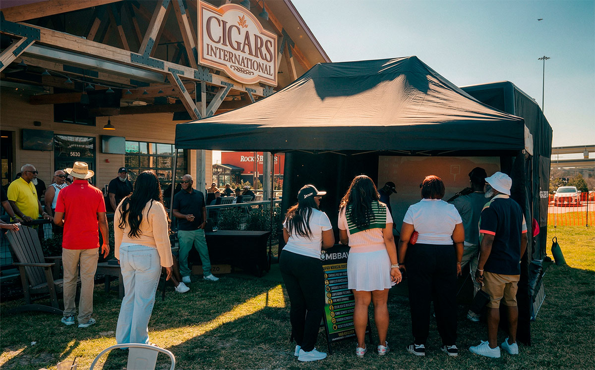 Group of people gathered outdoors near a black tent under a wooden sign reading 'Cigars International' during daytime.