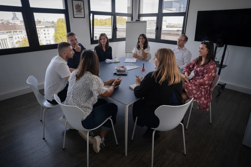 Groupe de huit personnes en réunion autour d'une table dans une salle de conférence lumineuse avec fenêtres offrant une vue sur la ville.