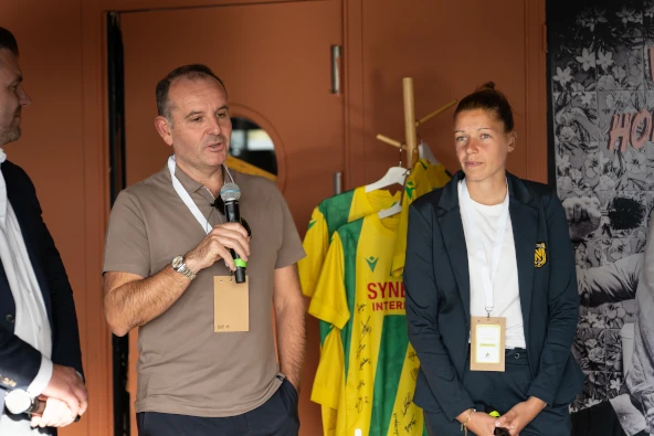 Un homme parlant au microphone à côté d'une femme en costume bleu, avec des maillots de football jaunes et verts suspendus en arrière-plan.