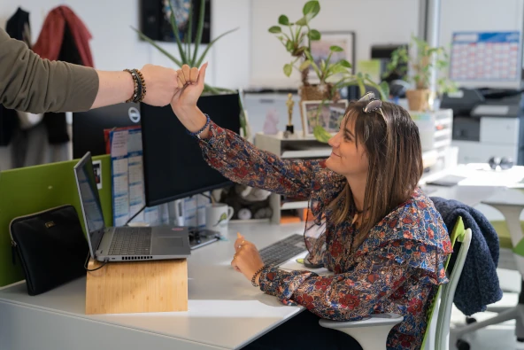 Une femme assise à un bureau fait un high five avec une autre personne dans un bureau lumineux avec des plantes et un ordinateur portable.