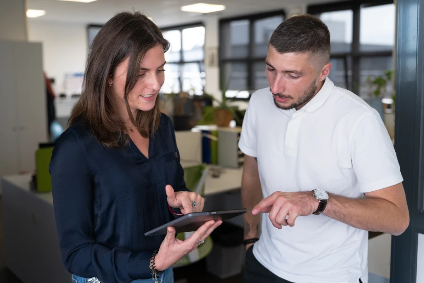 Deux collègues discutent en regardant et pointant une tablette dans un bureau moderne.