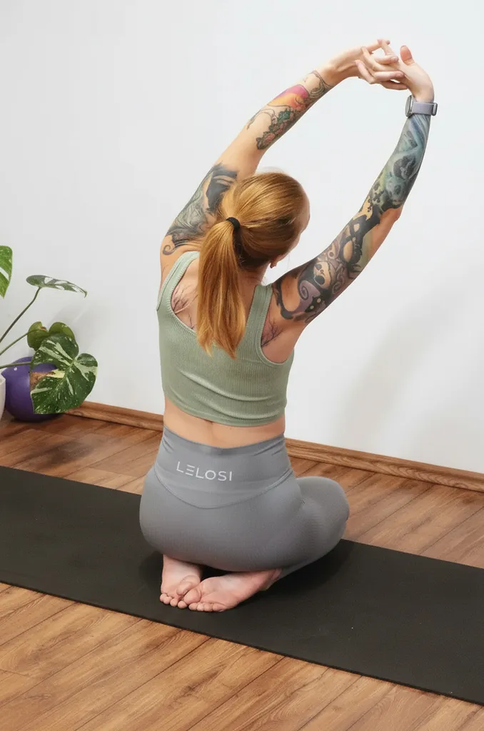 Žena s červenými vlasmi a tetovaniami sedí na jógovej podložke s rukami natiahnutými nad hlavou.