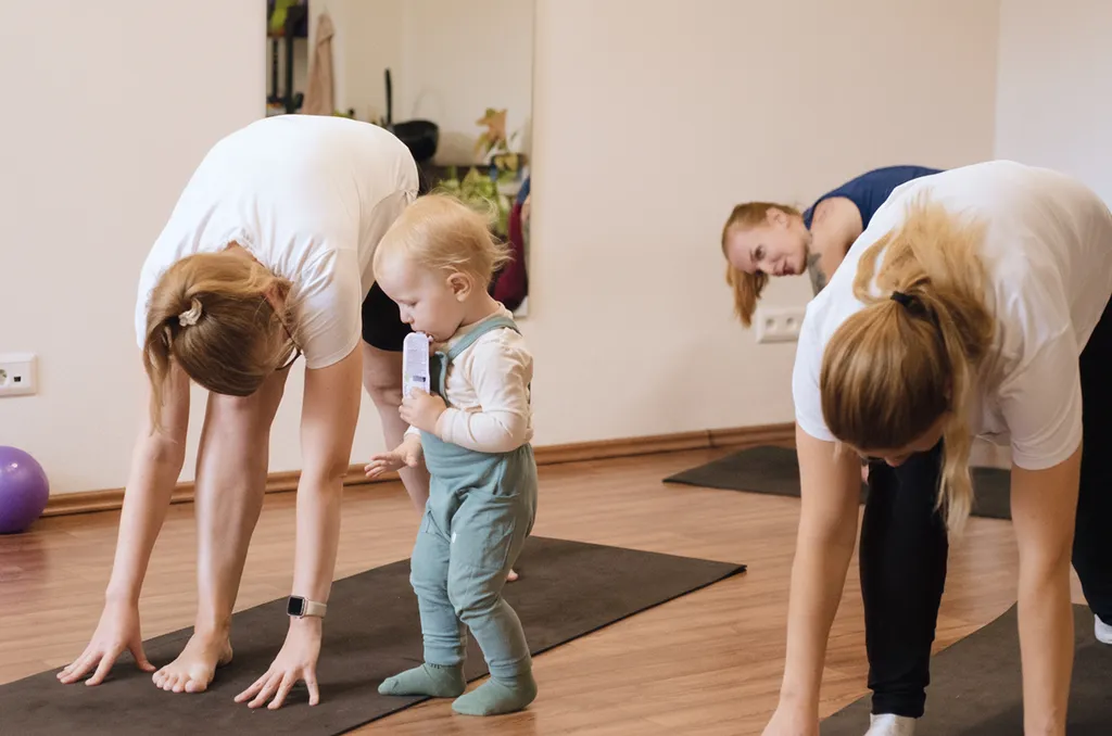 Dve ženy cvičia na jogových podložkách a malé dieťa stojí medzi nimi v miestnosti s drevenou podlahou.