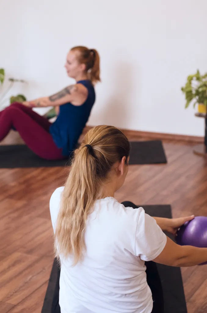 Dve ženy cvičiace na podložkách v interiéri, jedna drží fialový fitness loptu.