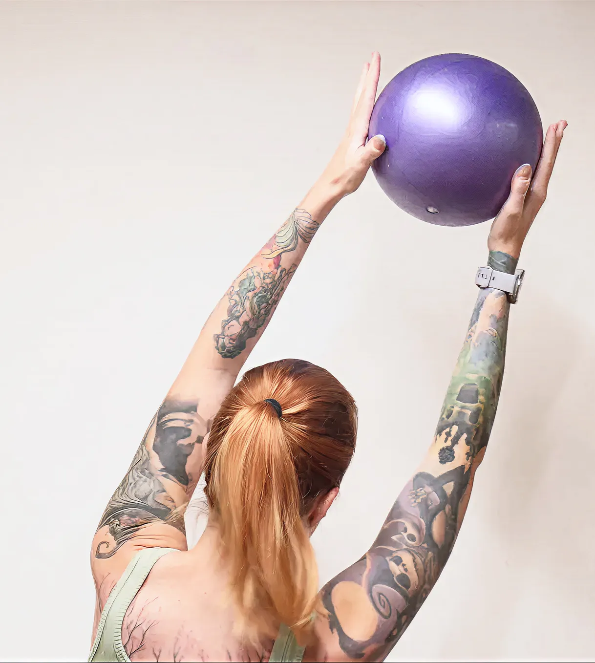 Žena s ryšavými vlasmi v copu drží nad hlavou fialový cvičebný loptu, má výrazné tetovania na pažiach.