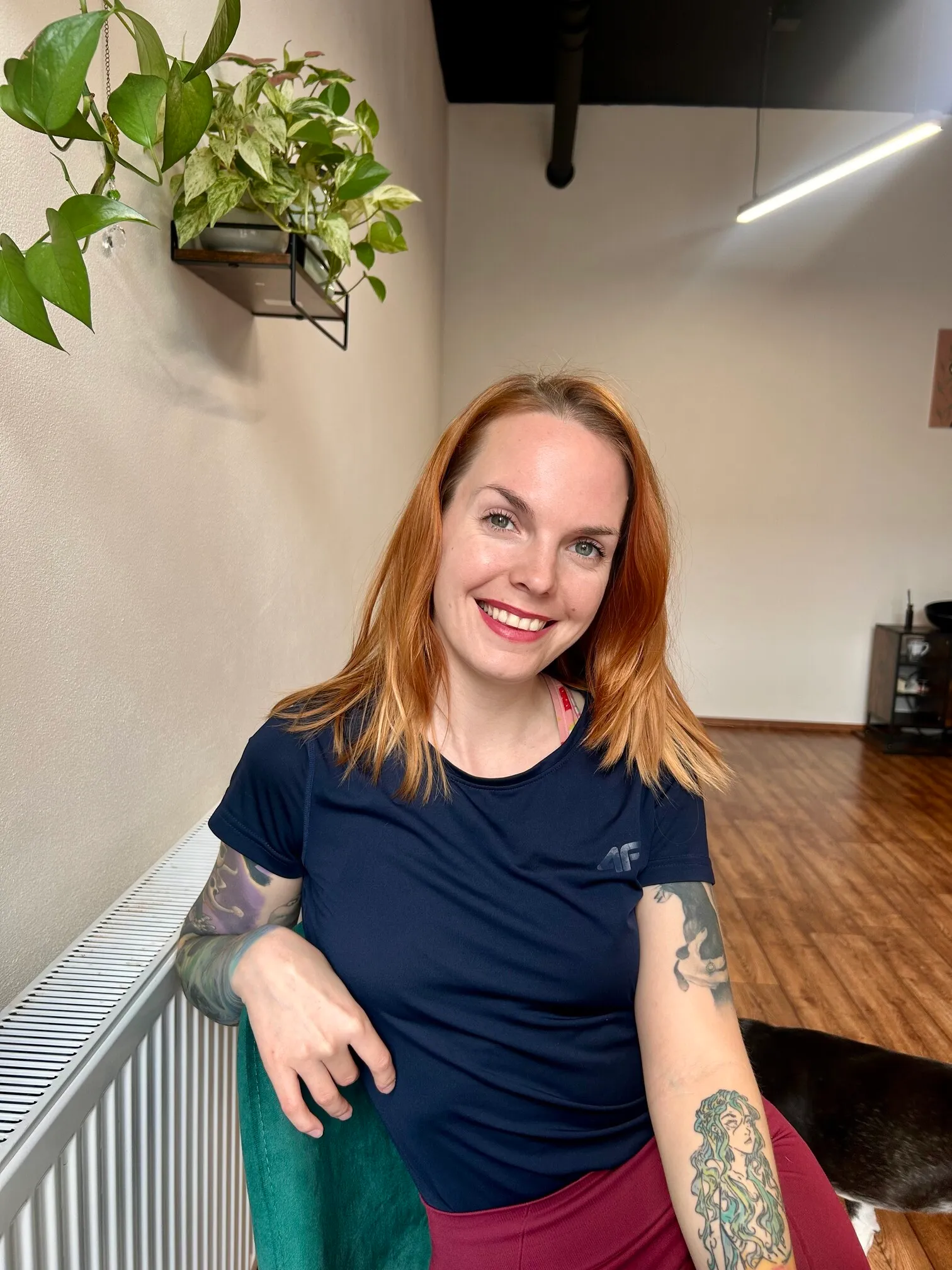 Usmievavá žena s červenými vlasmi a tetovaním na ruke sediaca na zelenej stoličke v izbe s drevenou podlahou a zelenou izbovou rastlinou na stene.