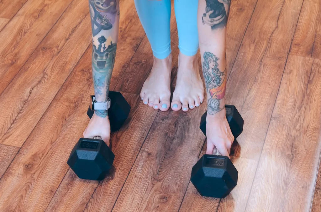 Osoba s farebnými tetovaniami na rukách drží činky na drevenej podlahe, vedľa sú bosé nohy s modrou farbou na nechtoch.