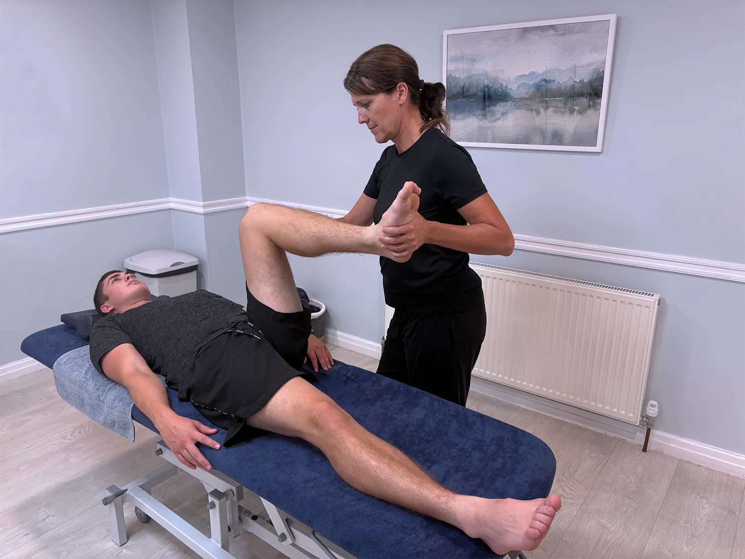 A physical therapist wearing black stretches the bent leg of a male patient lying on a treatment table in a clinical room with light blue walls and a framed landscape painting.