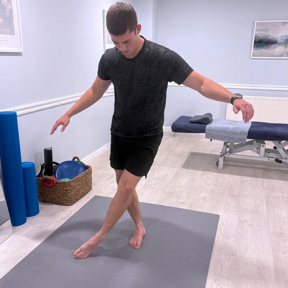 Young man in black shirt and shorts balancing barefoot on a gray exercise mat in a fitness or therapy room with exercise equipment and a treatment table.