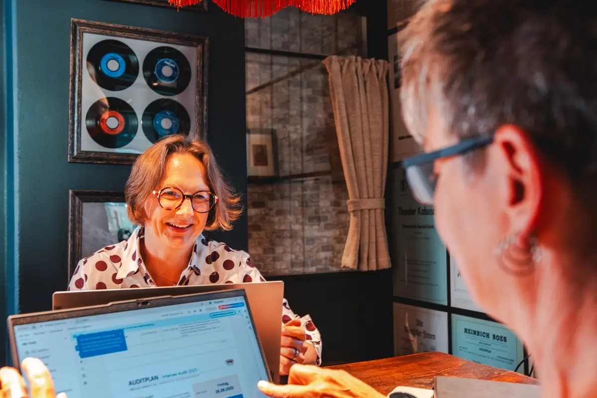 Zwei Frauen sitzen an einem Holztisch und unterhalten sich, eine zeigt auf einem Laptop-Bildschirm einen Aktionsplan.