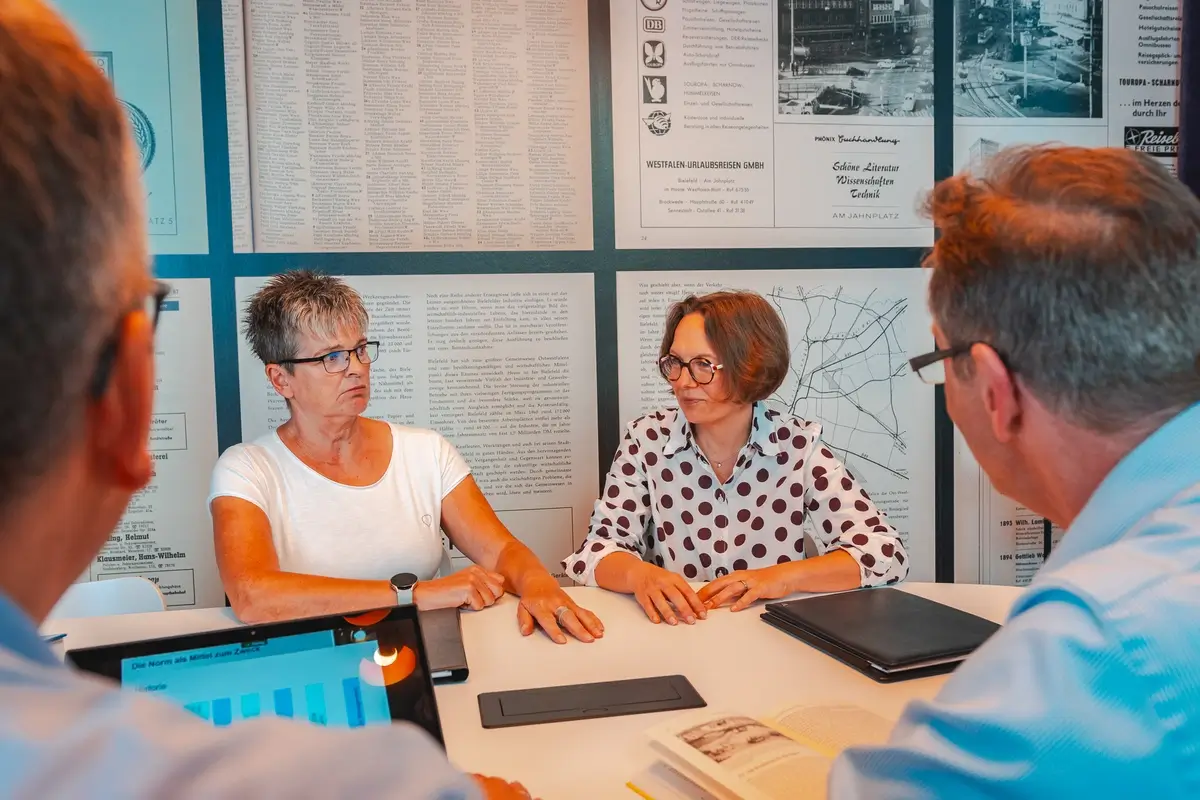 Vier Personen sitzen an einem runden Tisch in einem Gespräch, zwei Frauen mittleren Alters vorne, zwei Männer mit Brillen teilweise sichtbar.