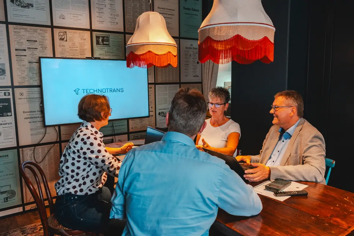 Vier Personen sitzen an einem Tisch in einem Besprechungsraum vor einem Bildschirm mit der Aufschrift TECHNOTRANS.