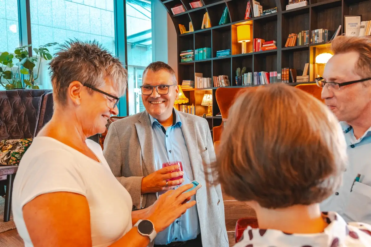 Vier Erwachsene unterhalten sich in einem gemütlichen Raum mit Büchernregalen im Hintergrund, eine Frau zeigt auf ihr Smartphone.