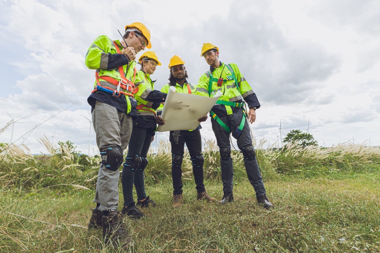 Vier Ingenieure mit Schutzhelmen und Sicherheitsausrüstung betrachten gemeinsam einen Plan auf einer grünen Wiese.