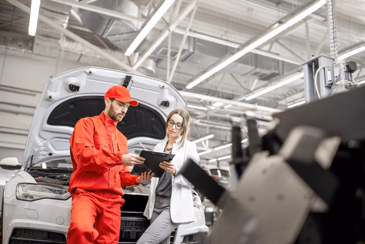 Ein Automechaniker in rotem Overall erklärt einer Frau mit Brille und weißen Blazer etwas auf einem Klemmbrett vor einem Auto mit geöffneter Motorhaube in einer Werkstatt.