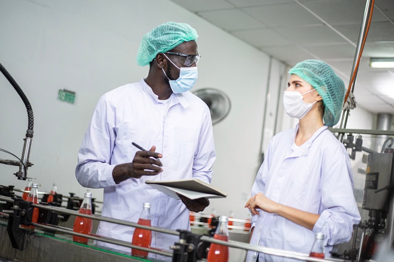 Zwei Fabrikarbeiter in Schutzkleidung und Masken überwachen Flaschen mit rotem Getränk auf einer Produktionslinie.