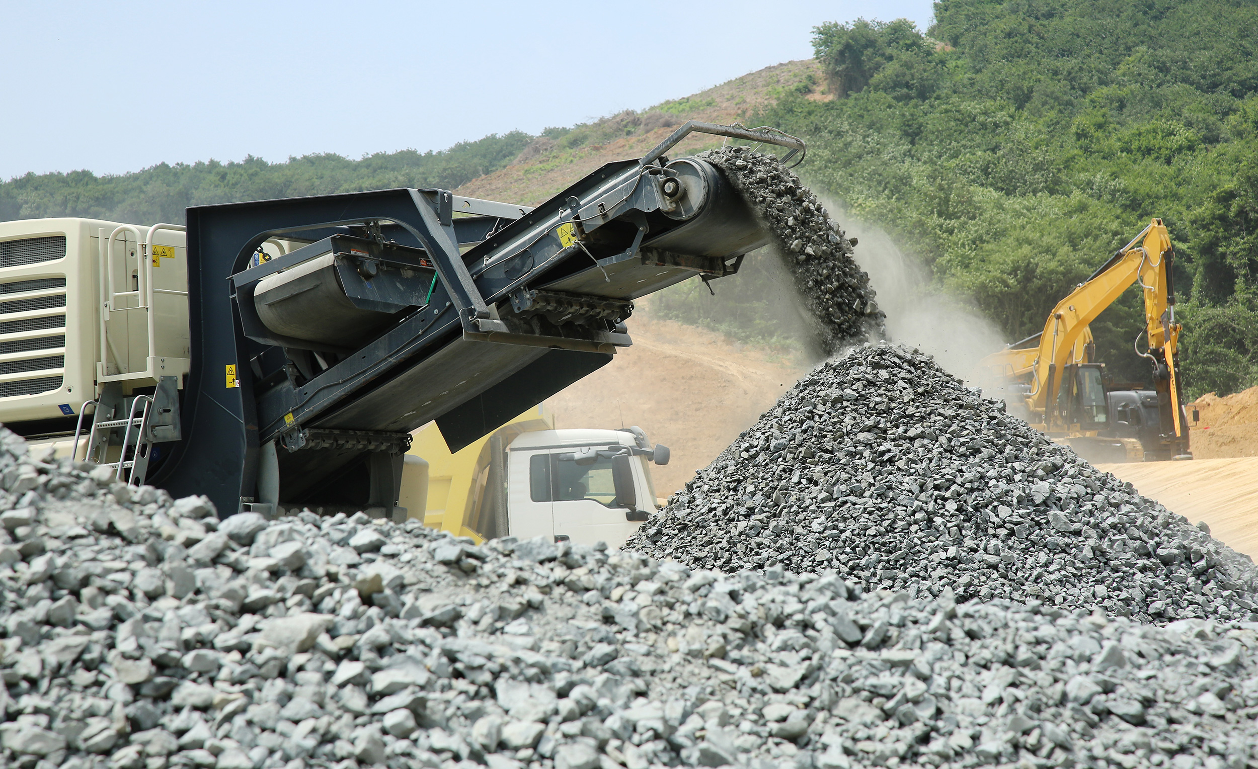 Conveyor belt depositing crushed rocks into a large pile at a construction site with excavator and truck in the background.