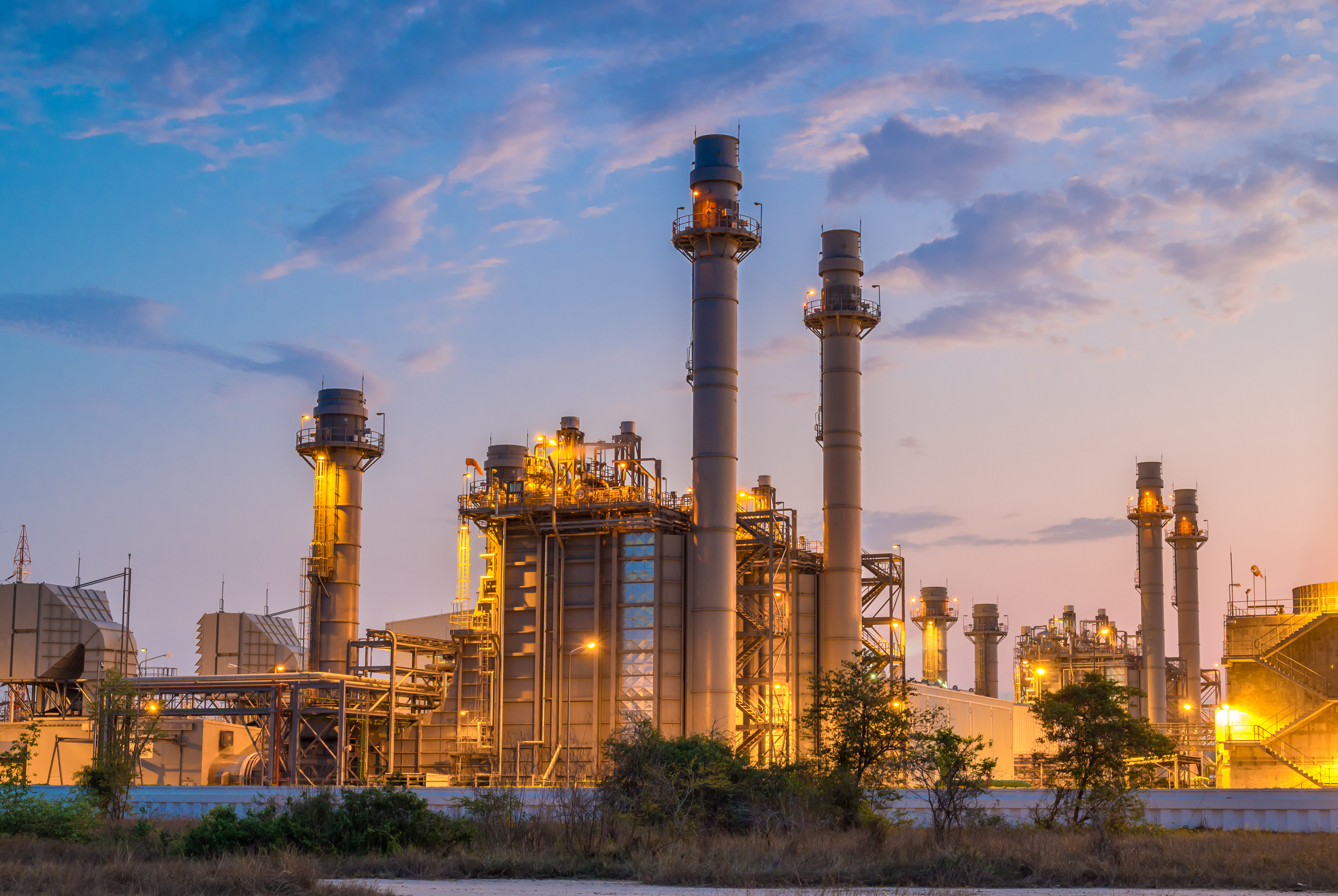 Power plant with tall exhaust stacks lit by yellow lights at dusk under a partly cloudy sky.