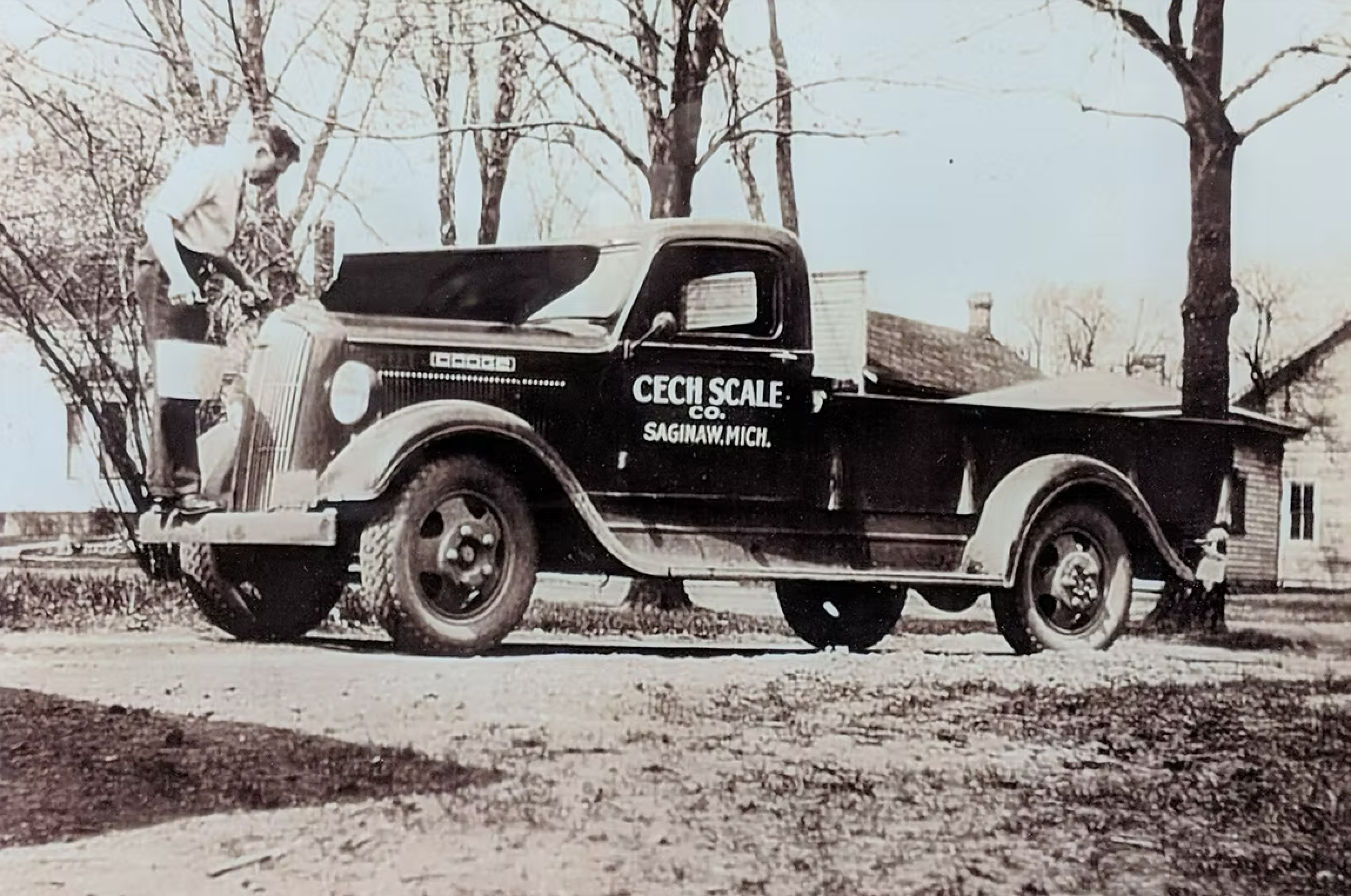 Vintage black pickup truck with open hood and company name 'CECH SCALE CO. SAGINAW, MICH.' on the door, with a man inspecting the engine.