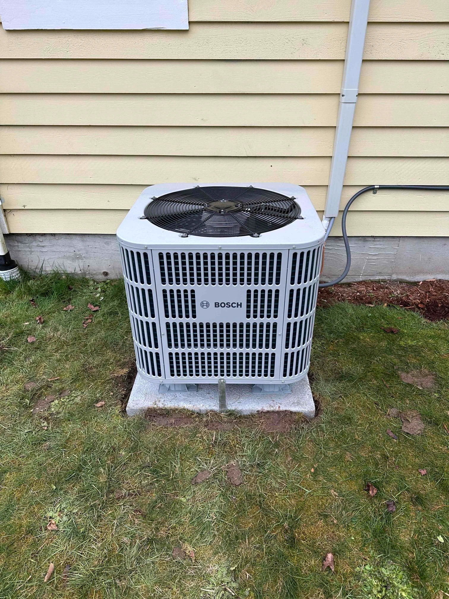 A gray Bosch air conditioner unit is mounted on a concrete platform beside a yellow house with horizontal siding,