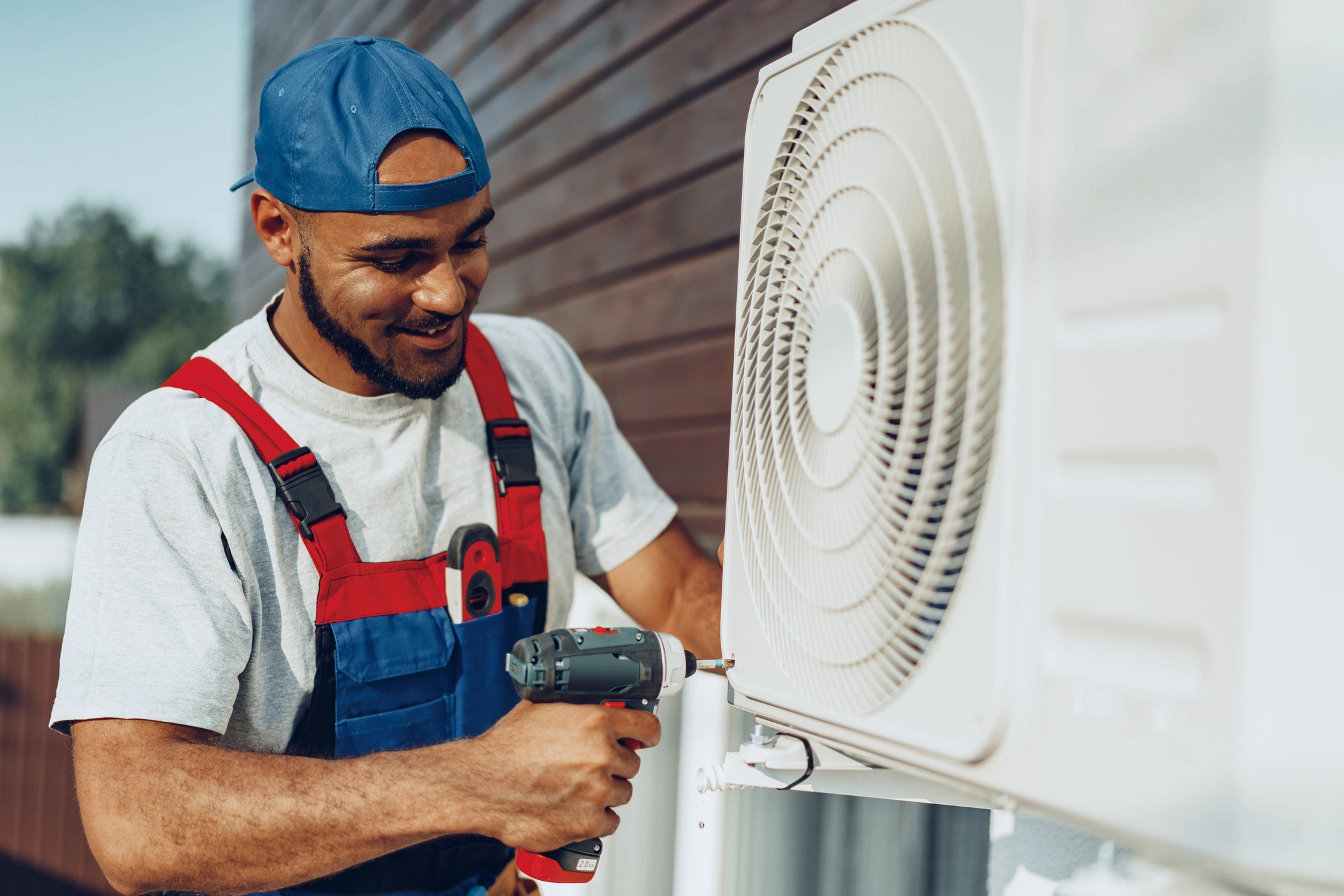 Man in blue cap and red overalls smiling while fixing an air conditioning unit with a power drill.