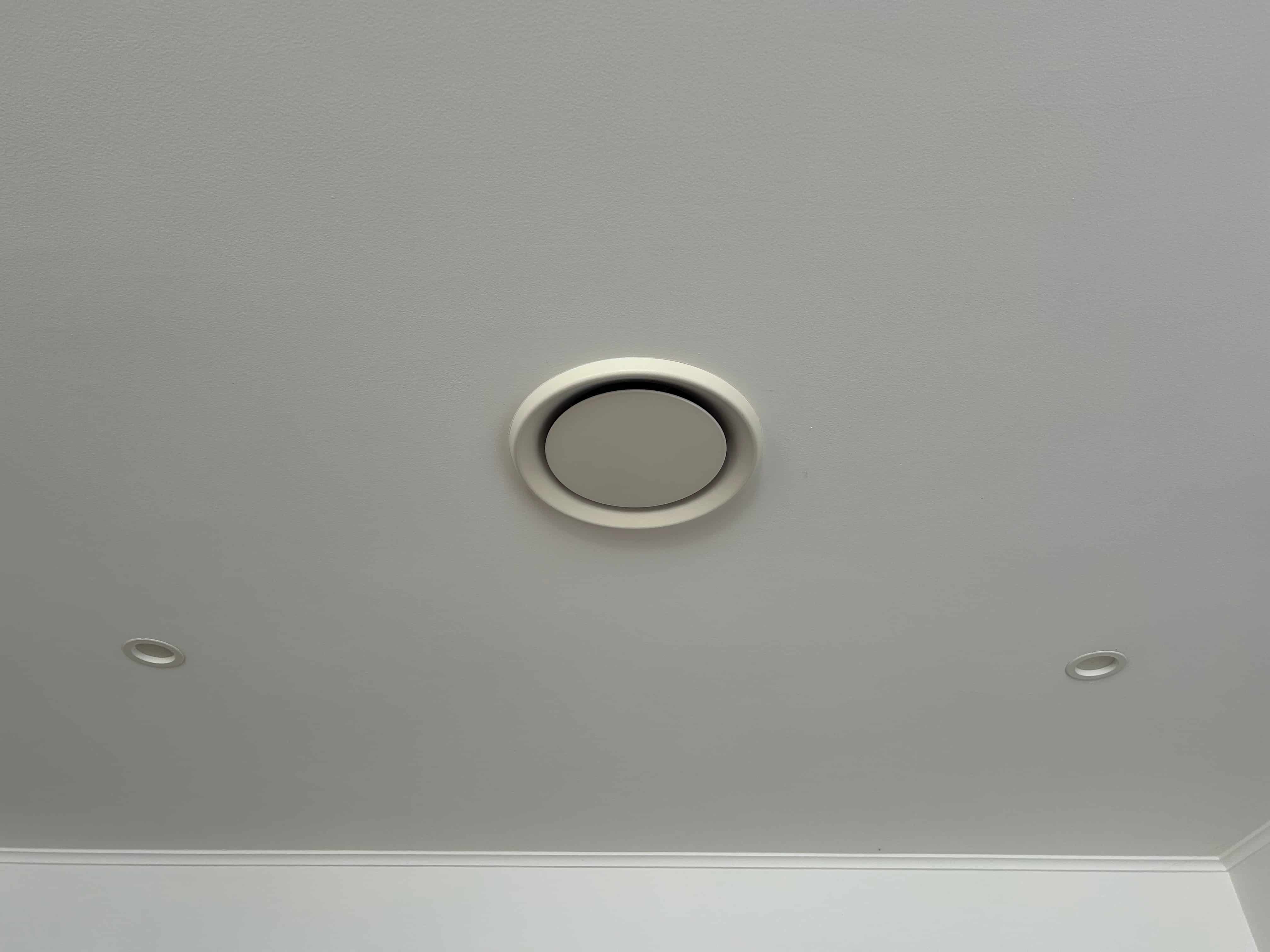 Ceiling-mounted round ventilation diffuser centered between two recessed lights in a modern room.