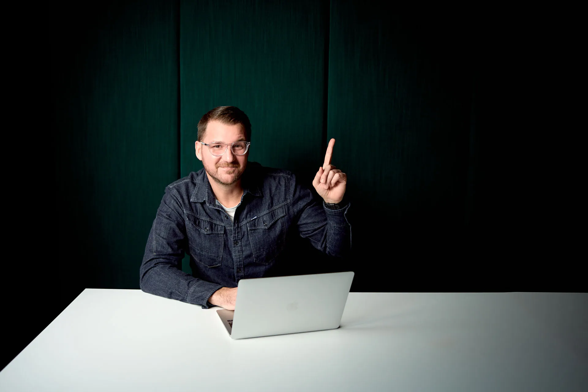 Philip Fuchslocher am Schreibtisch mit Laptop sitzend, während er in die Kamera lächelt und den Zeigefinger hebt.