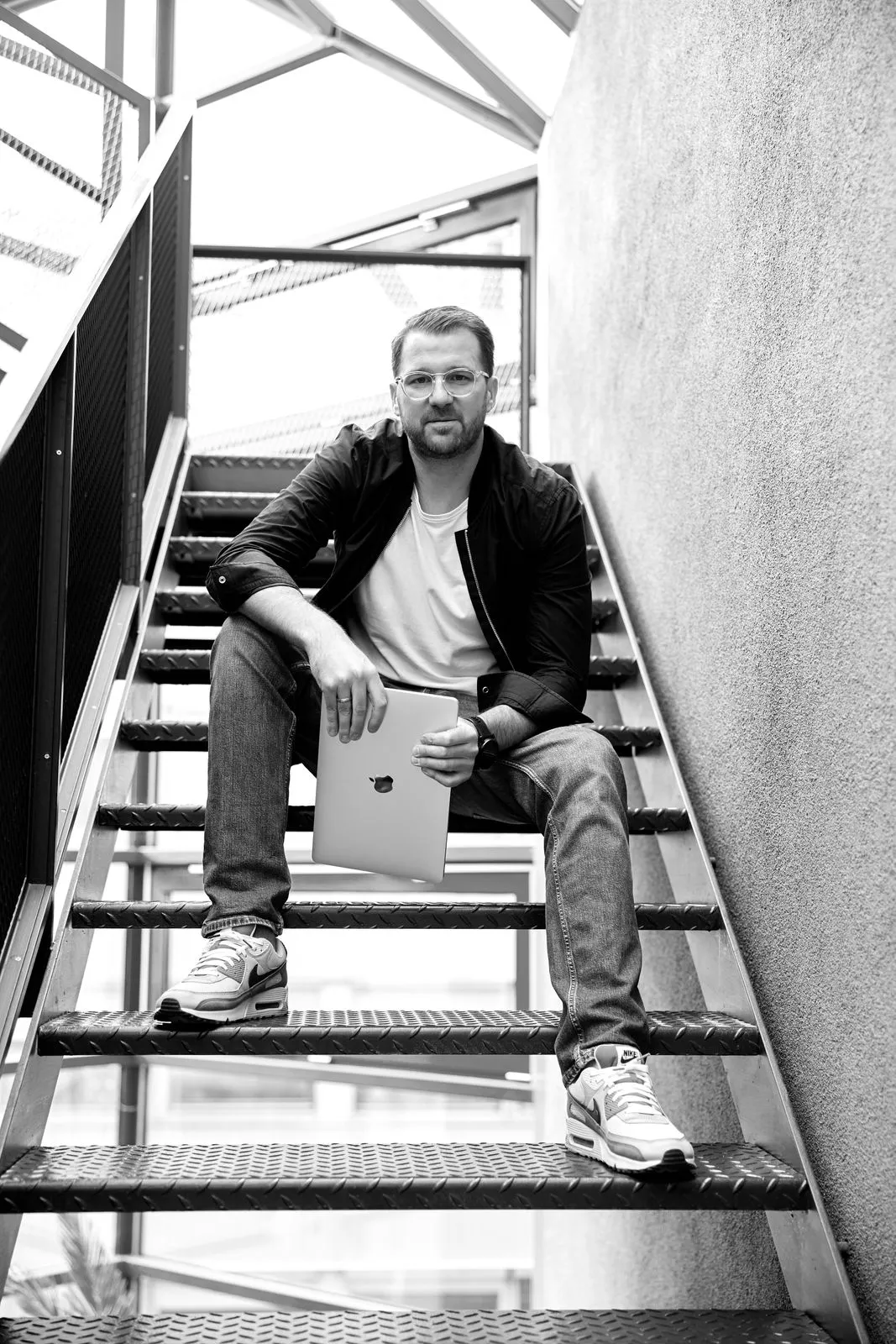 Philip Fuchslocher sitzt auf einer Treppe und hält einen Laptop in der Hand, dabei lächelt er in die Kamera in Schwarz/Weiß-Optik