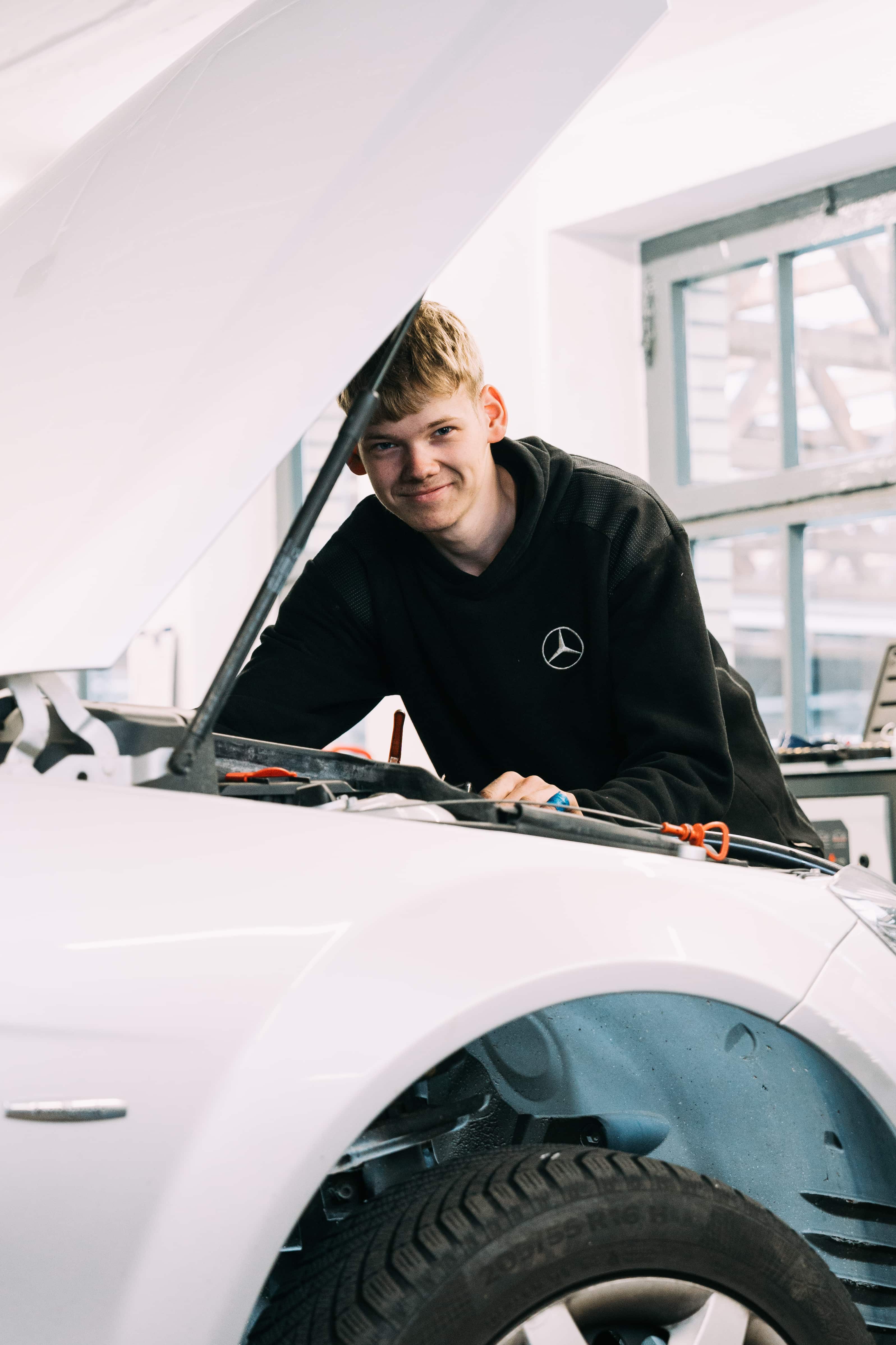 Auszubildender lehnt lächelnd über dem Motorraum eines Fahrzeugs