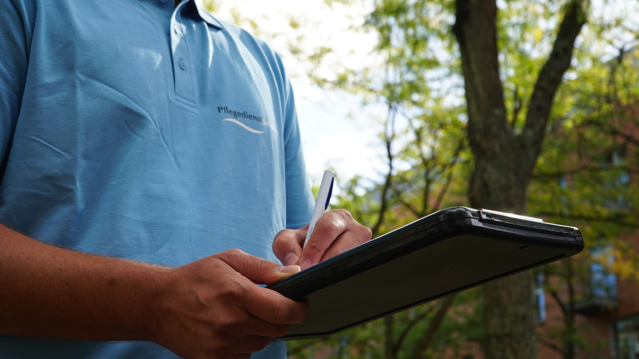 Person in blauem Poloshirt mit der Aufschrift ‚Pflegedienst‘ schreibt mit einem Stift auf einem Klemmbrett im Freien.