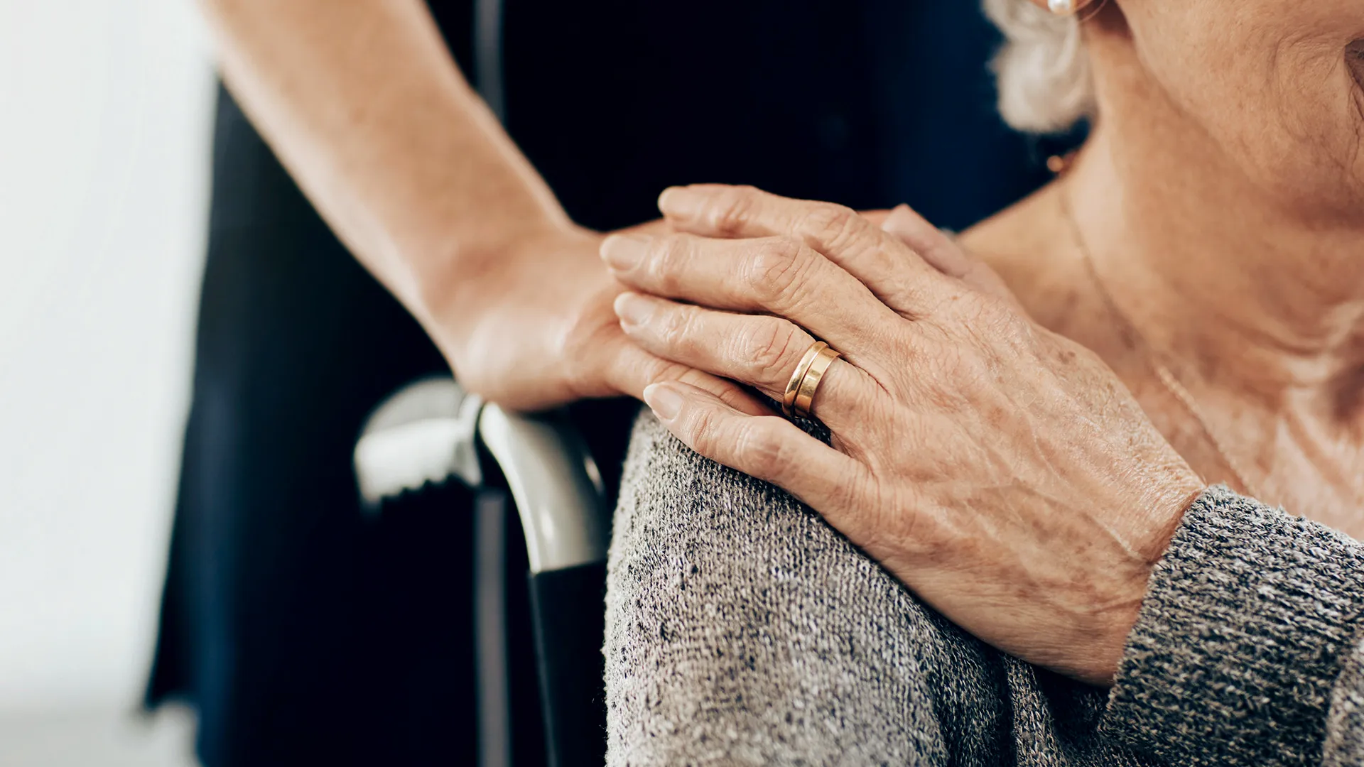Eine Hand legt sich fürsorglich auf die Schulter einer älteren Frau mit grauem Pullover.