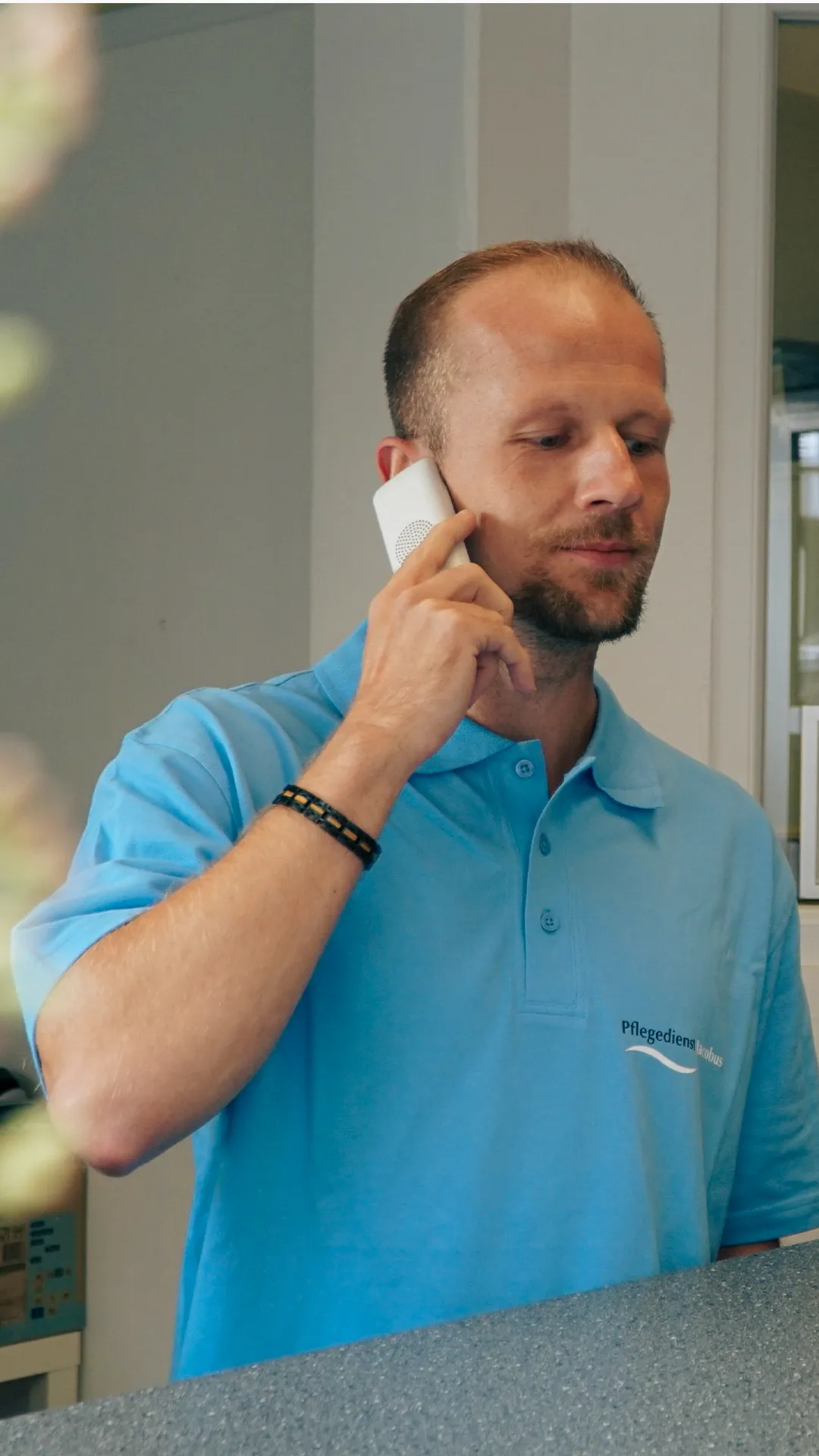 Mann in blauem Poloshirt mit Schriftzug „Pflegedienst Jacobus“ telefoniert am Empfang.