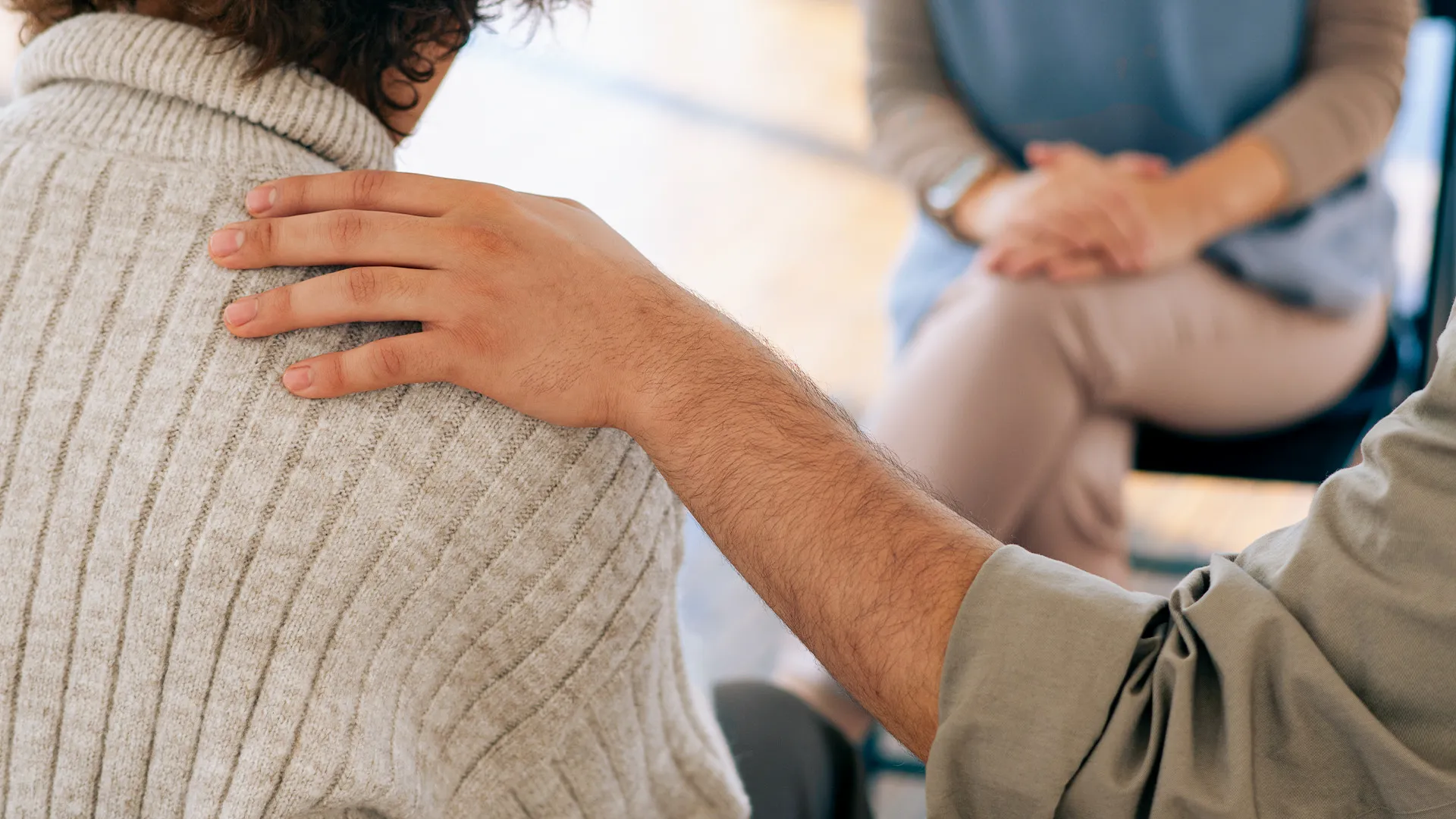 Eine Person legt tröstend die Hand auf die Schulter einer anderen Person, die einen beigen Pullover trägt, während eine dritte Person im Hintergrund sitzt.