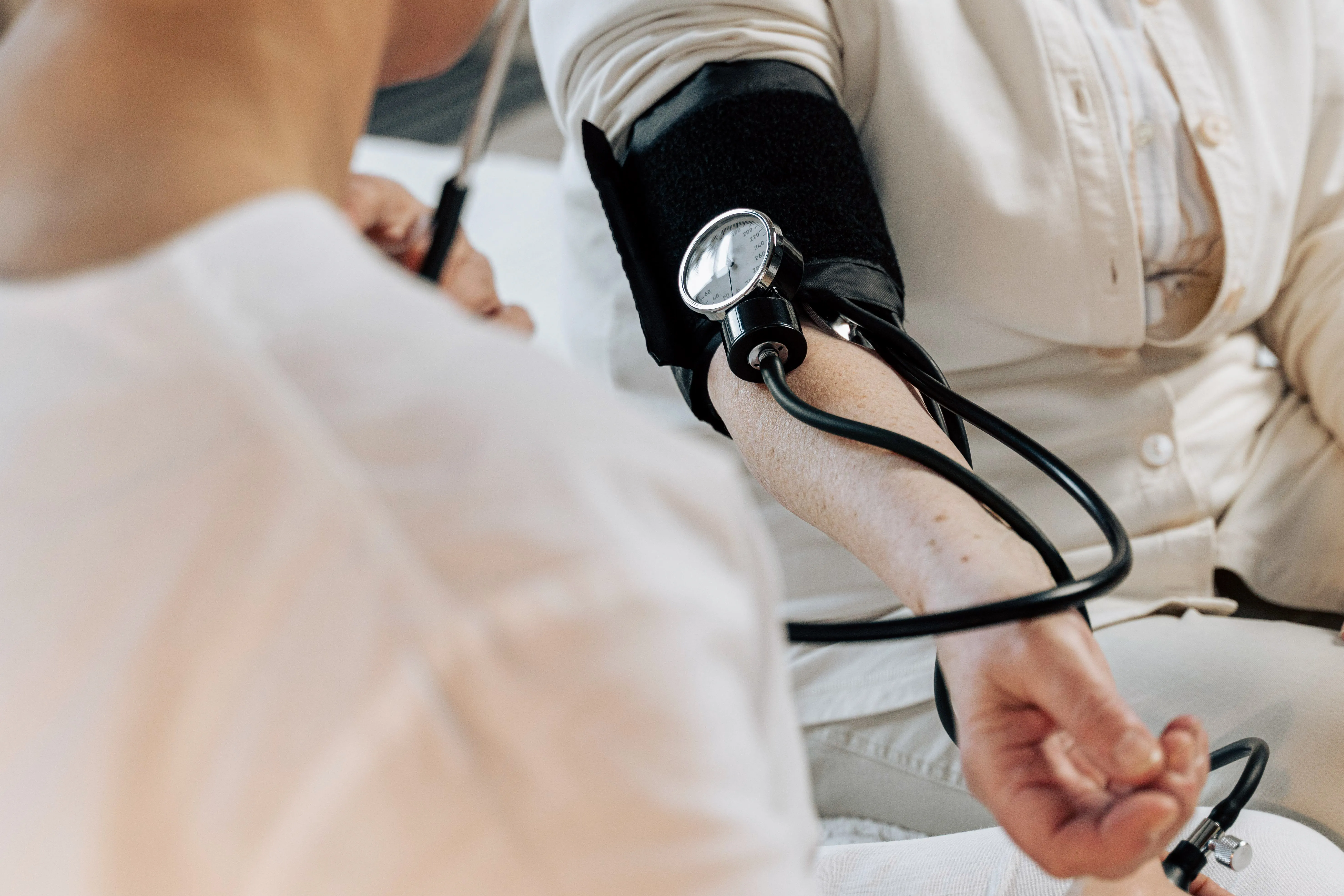 Person misst Blutdruck mit Manschette am linken Oberarm, betreut von einer anderen Person.