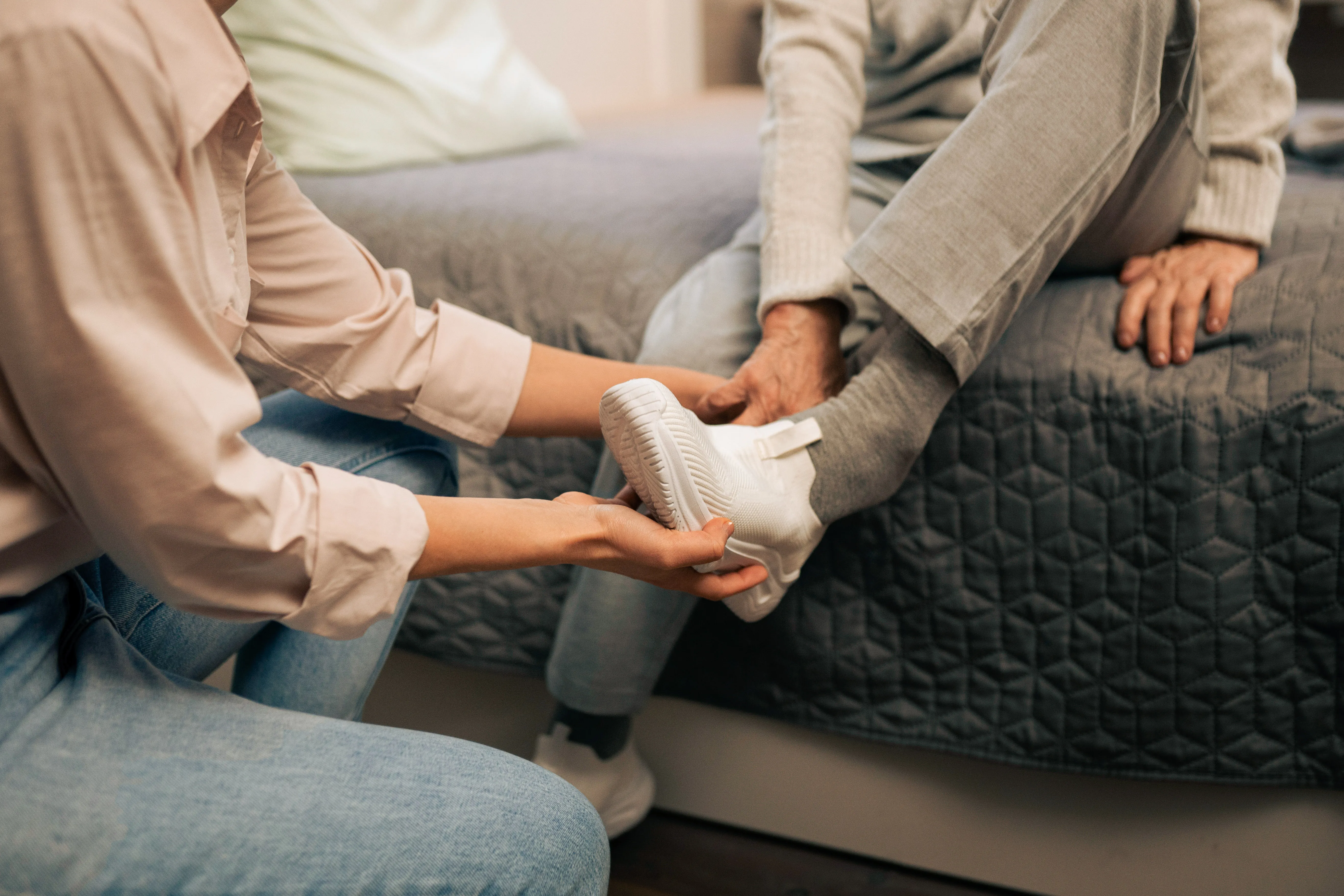 Pflegekraft hilft älterer Person, einen weißen Schuh anzuziehen, während diese auf einem Bett sitzt.