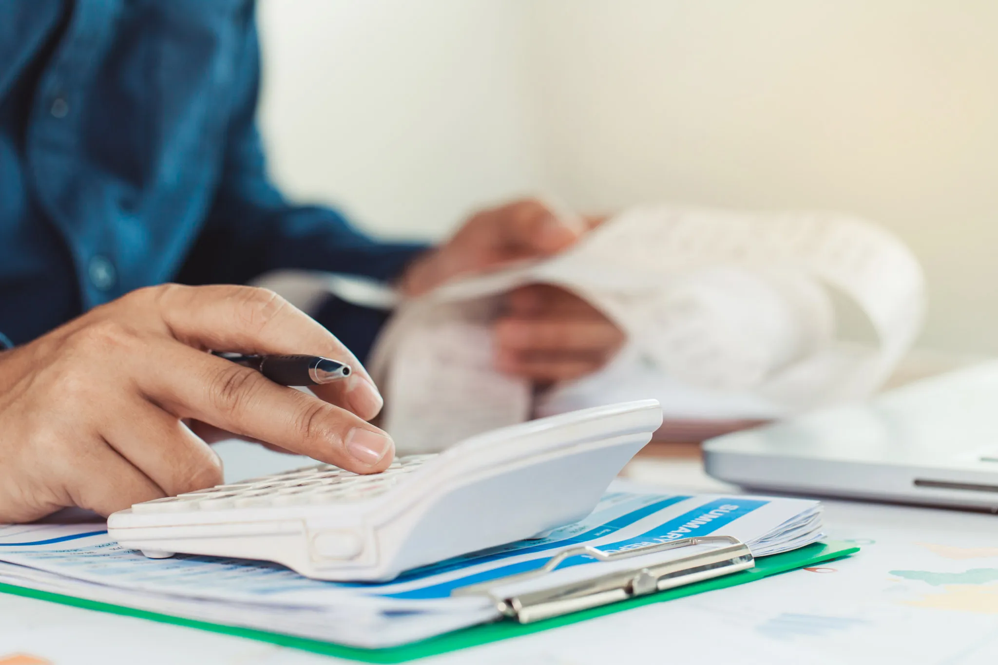 Person benutzt Taschenrechner, hält Stift und prüft Belege auf einem Schreibtisch mit Dokumenten und Laptop.