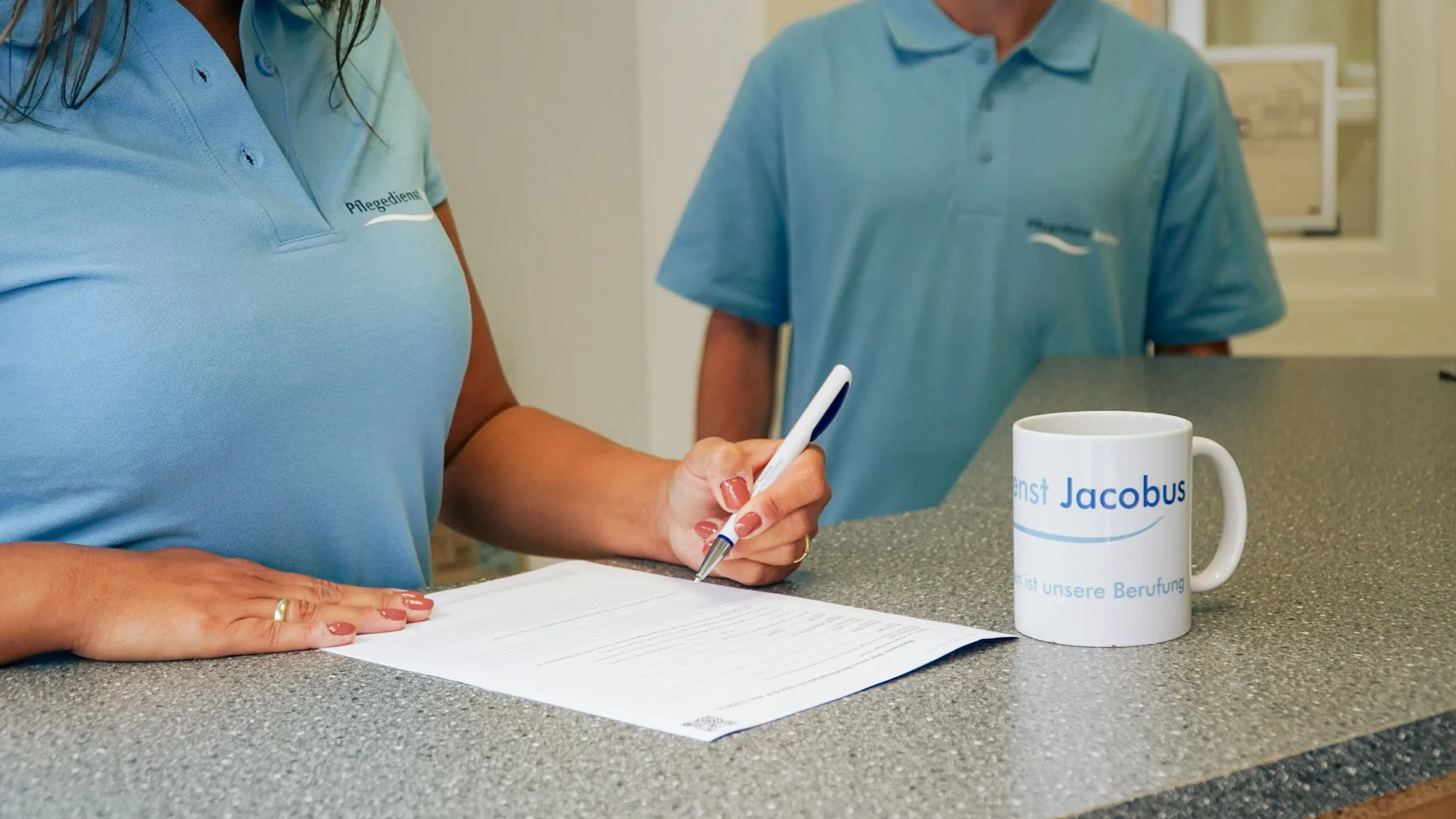 Zwei Personen in hellblauen Poloshirts an einem Tisch, eine Person unterschreibt ein Dokument, daneben steht eine Tasse mit der Aufschrift Pflegedienst Jacobus.