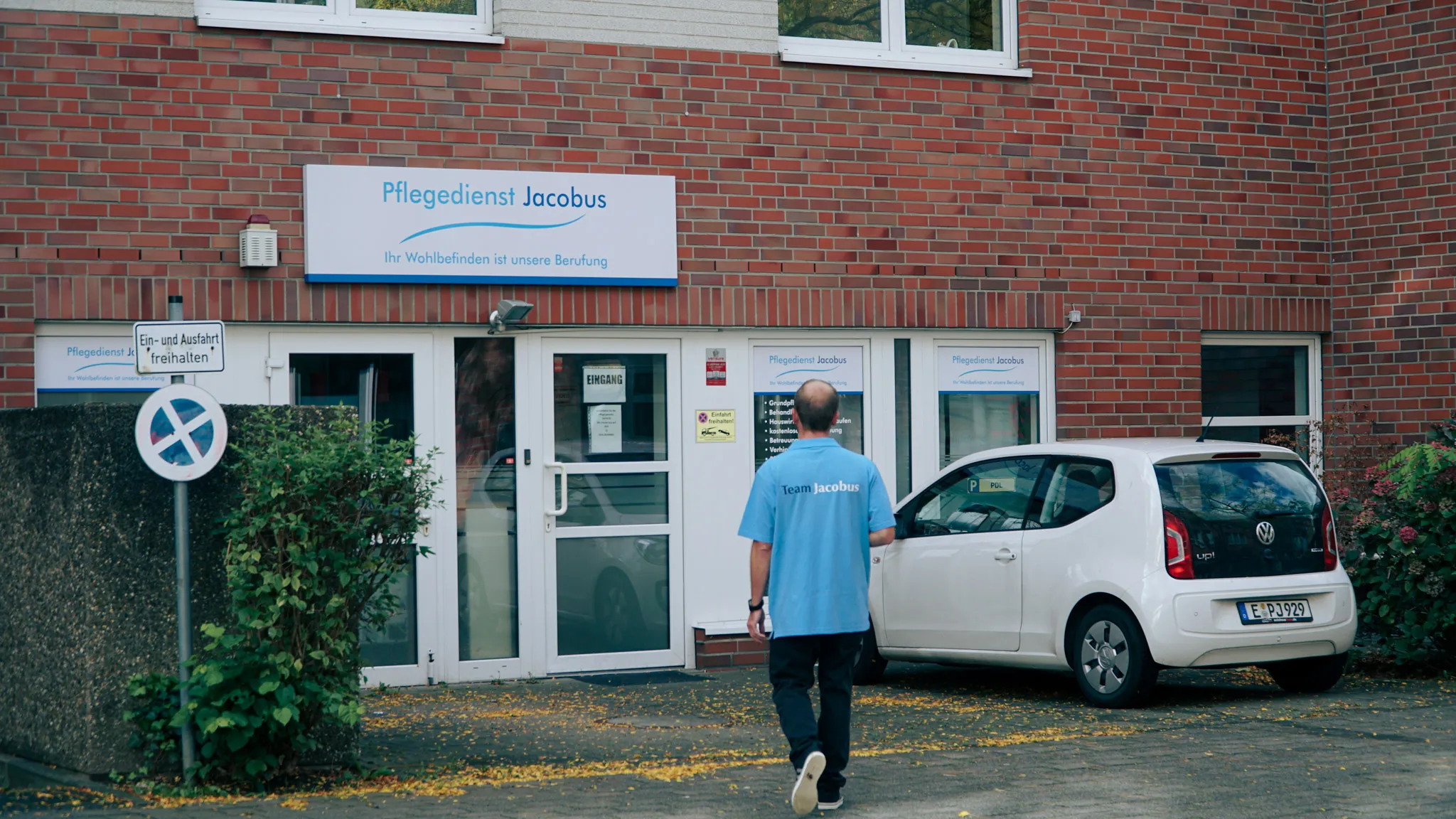 Mann mit blauem Team Jacobus T-Shirt geht auf den Eingang eines Gebäudes mit einem weißen VW Up, geparkt auf der rechten Seite.