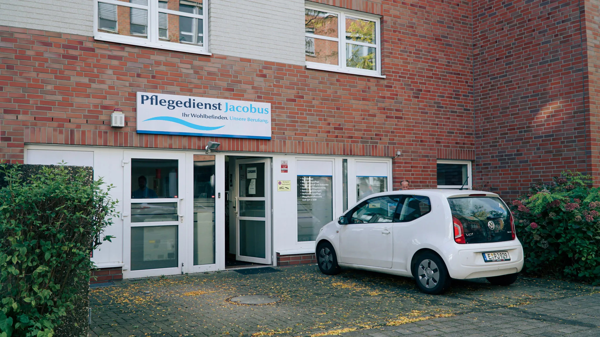 Eingang eines Backsteingebäudes mit Schild 'Pflegedienst Jacobus' über der Tür und einem geparkten weißen VW Up.