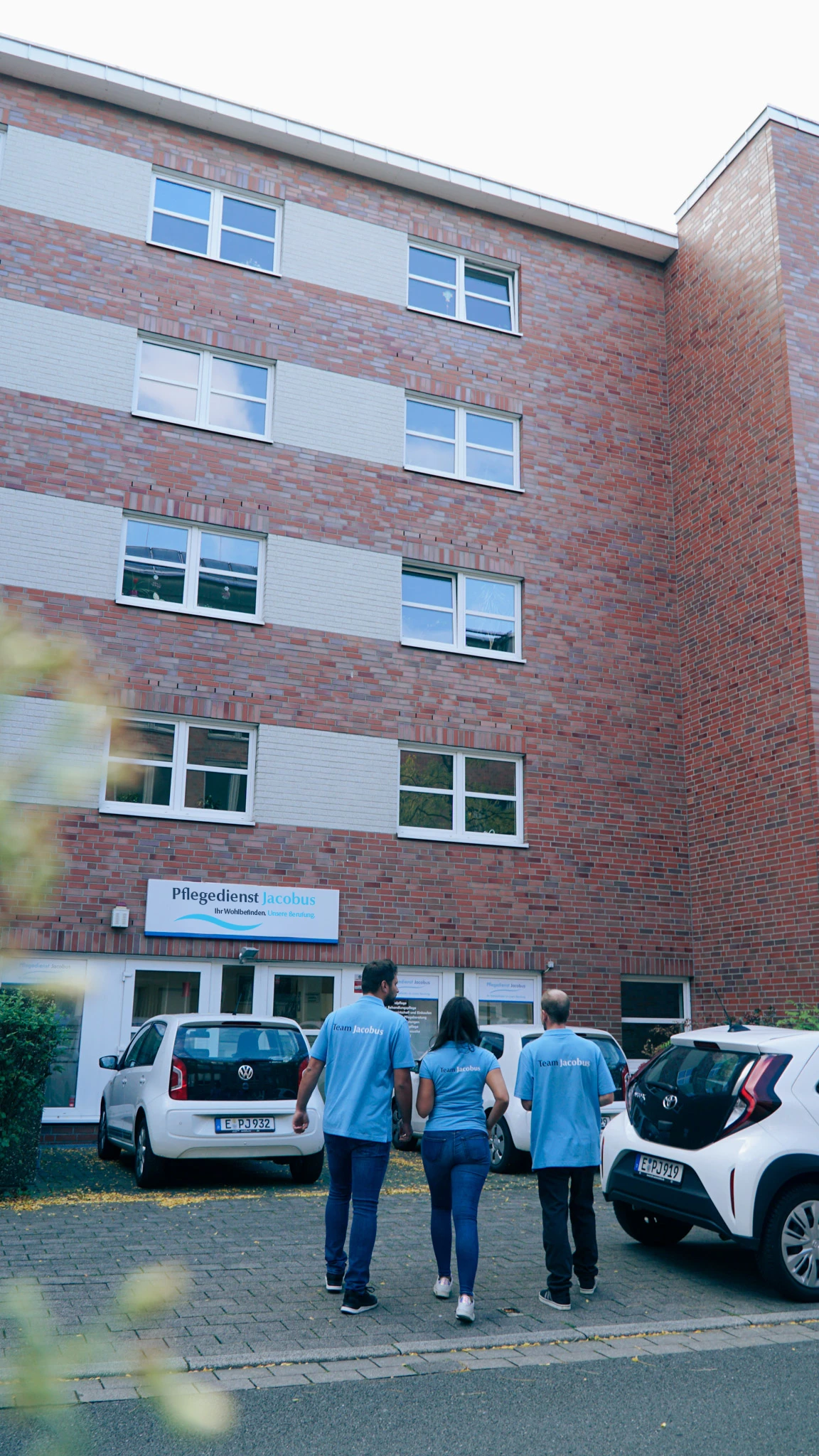 Drei Personen in blauen T-Shirts mit der Aufschrift „Team Jacobus“ gehen auf ein Gebäude mit Schild „Pflegedienst Jacobus“ zu, zwei geparkte Autos stehen davor.