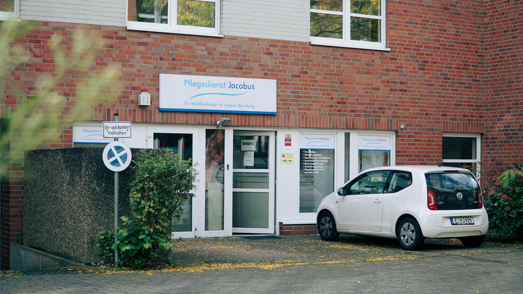 Eingang eines Pflegedienstes mit weißen Türen, Schild 'Pflegedienst Jacobus' und einem weißen VW Up! Fahrzeug davor.