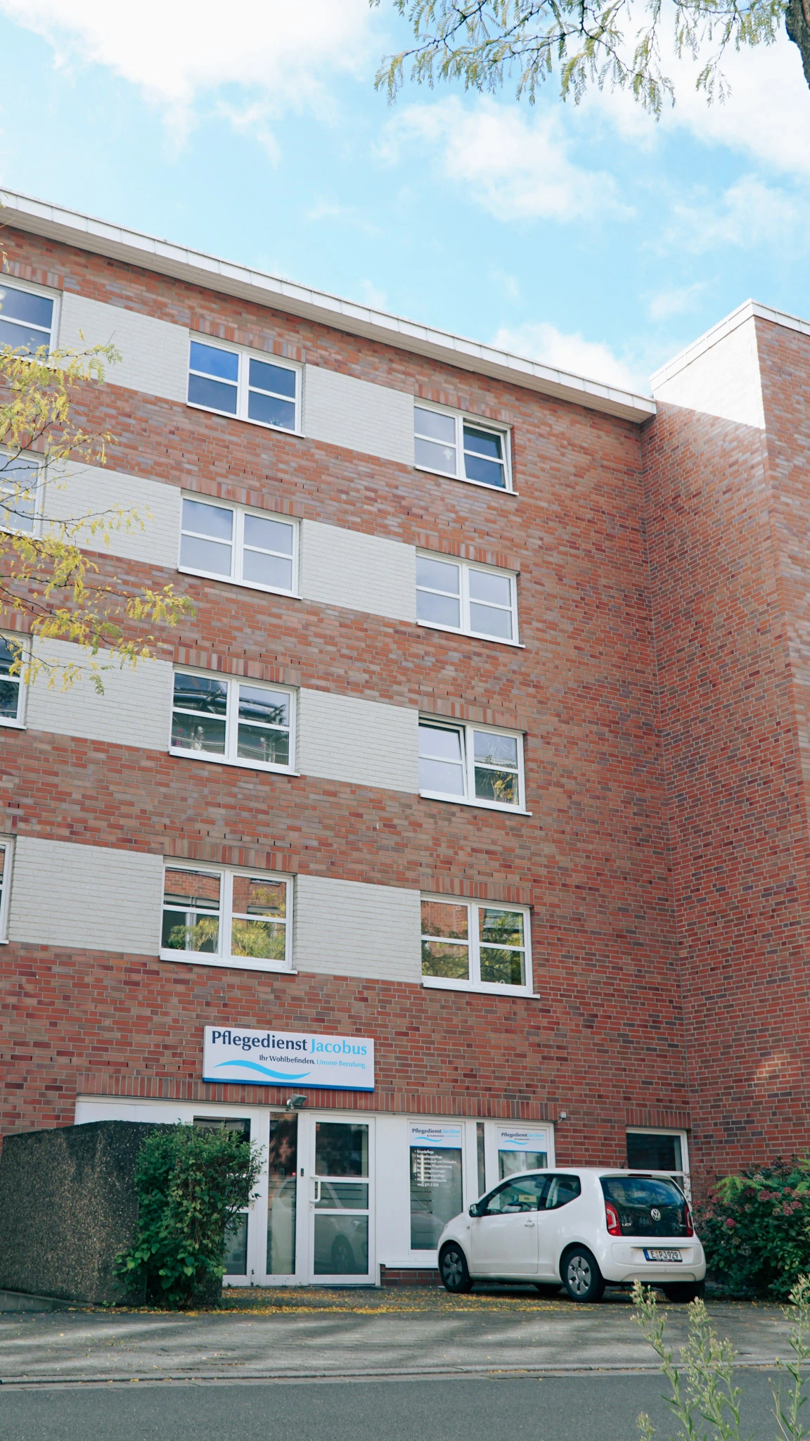 Gebäude mit rotem Backstein und weißen Fensterrahmen, davor ein geparkter weißer Volkswagen und ein Schild mit der Aufschrift 'Pflegedienst Jacobus'.