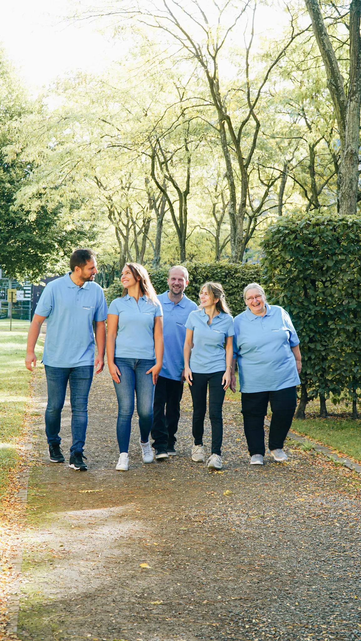 Fünf Personen in hellblauen Poloshirts laufen lächelnd auf einem von Bäumen gesäumten Weg im Park.